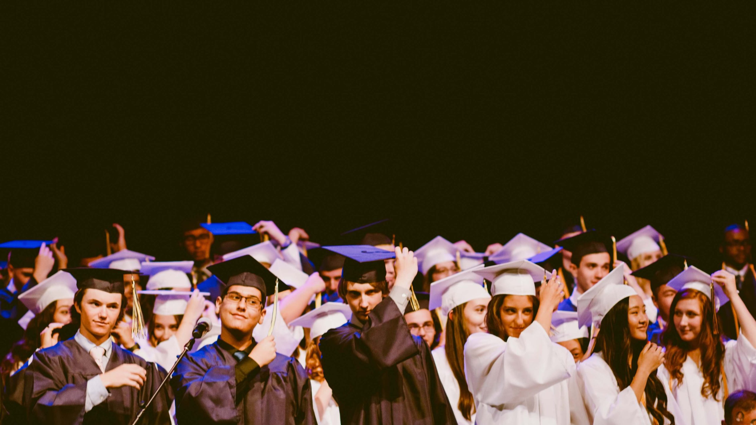 A large group of students dressed in black and white graduation gowns and caps celebrating their commencement, moving their tassels to the side in a traditional graduation gesture on stage.