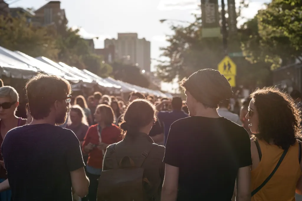 A diverse group of people walking through a lively outdoor street festival, with tents, vendors, and a bustling crowd in the background, illuminated by the warm glow of the setting sun.