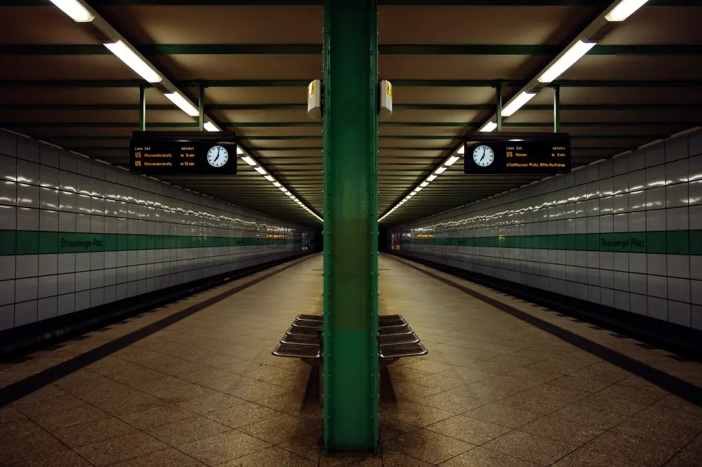 A perfectly symmetrical view of the empty Strausberger Platz subway station, featuring a central green pillar, tiled walls, overhead lighting, and digital departure boards displaying train schedules.