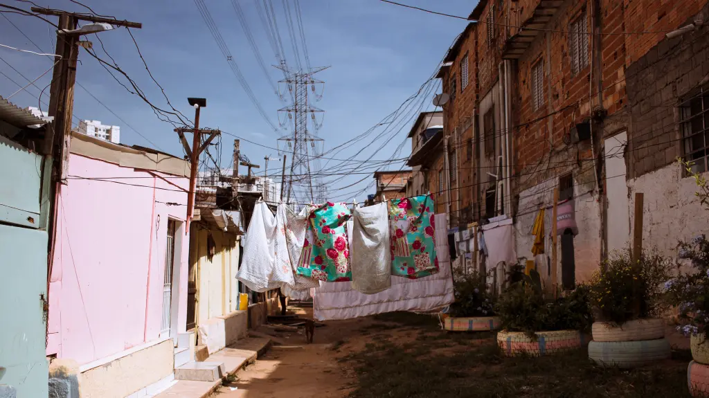 A narrow dirt alleyway in a modest urban neighborhood with colorful clothes hanging on a clothesline between brick and pastel-painted houses, surrounded by electrical wires and potted plants made from repurposed tires.