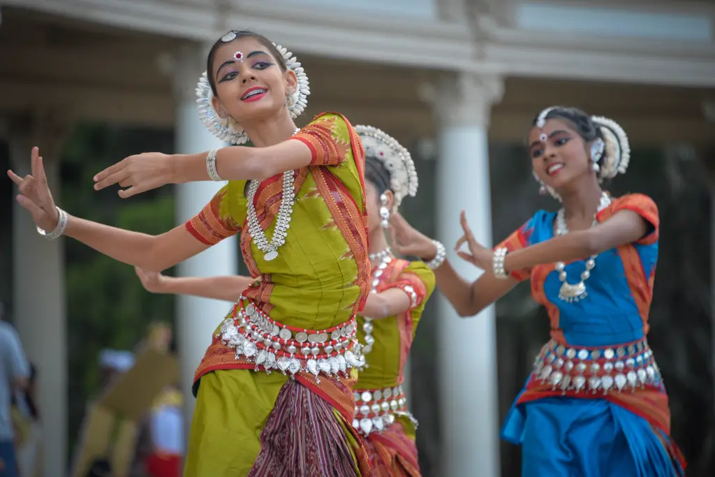 A group of young women dressed in vibrant traditional Indian attire gracefully performing the classical Odissi dance, adorned with intricate jewelry and expressive facial expressions in an outdoor cultural setting.