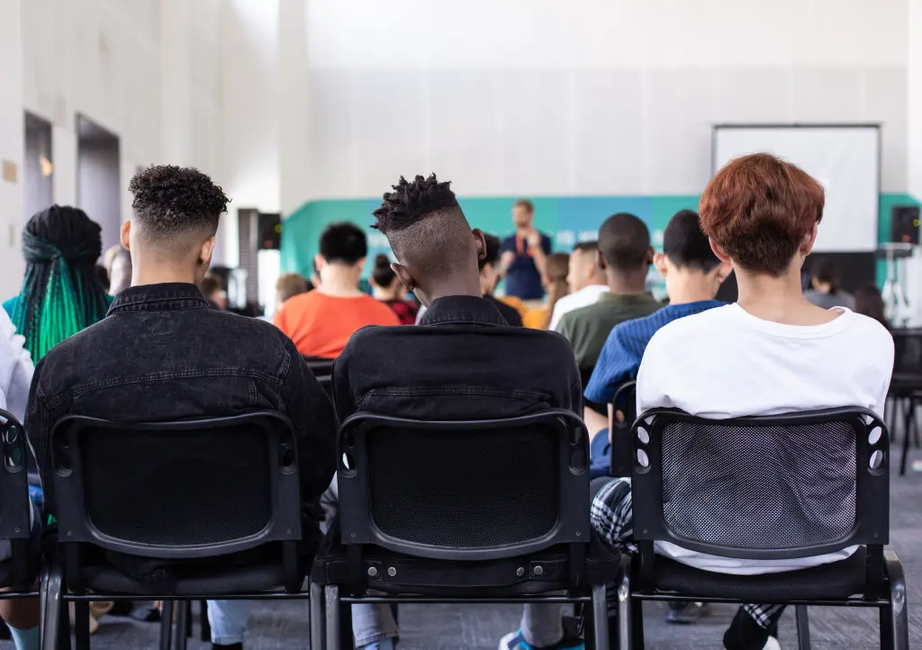 A group of young people sitting in black chairs, attentively listening to a speaker at the front of a modern conference room with a projector screen in the background