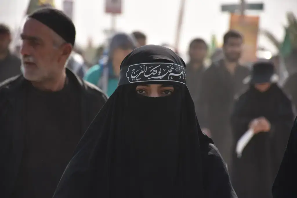 A woman wearing a black niqab and a headband with Arabic script stands among a crowd of people dressed in black, some carrying flags, in what appears to be a public or religious procession.