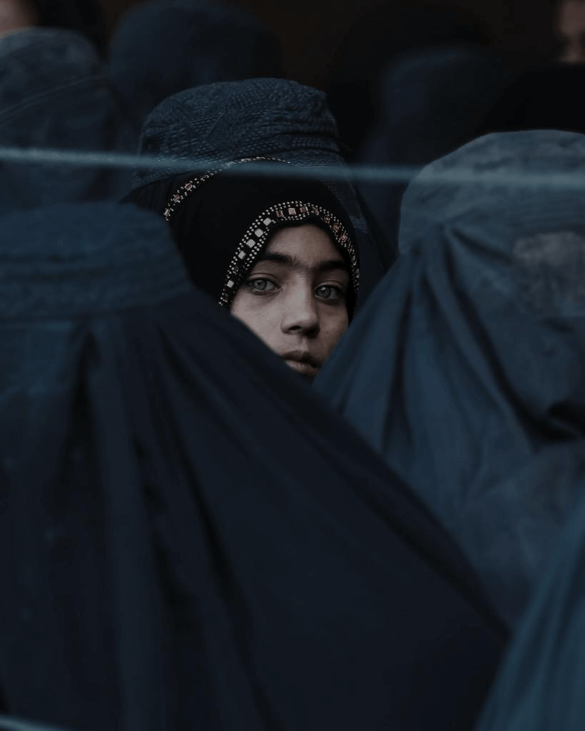 A young woman wearing a black hijab with golden details gazes directly at the camera, surrounded by others dressed in dark blue veils, creating a mysterious and poignant atmosphere.