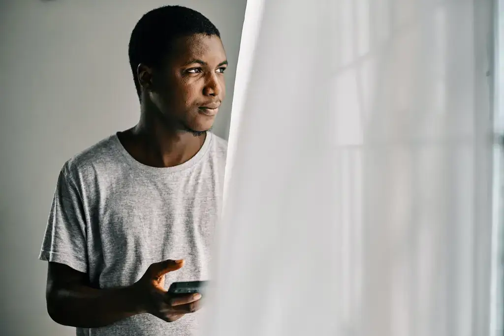 A man in a gray t-shirt holding a smartphone, gazing thoughtfully out of a bright window with soft white curtains.