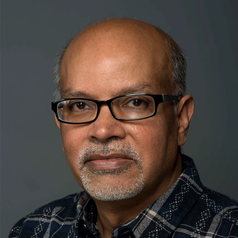 A professional portrait of Imran Omer, the author of 'Entangled Lives.' He is wearing glasses and a dark blue checkered shirt, with a neutral expression and a gray background.